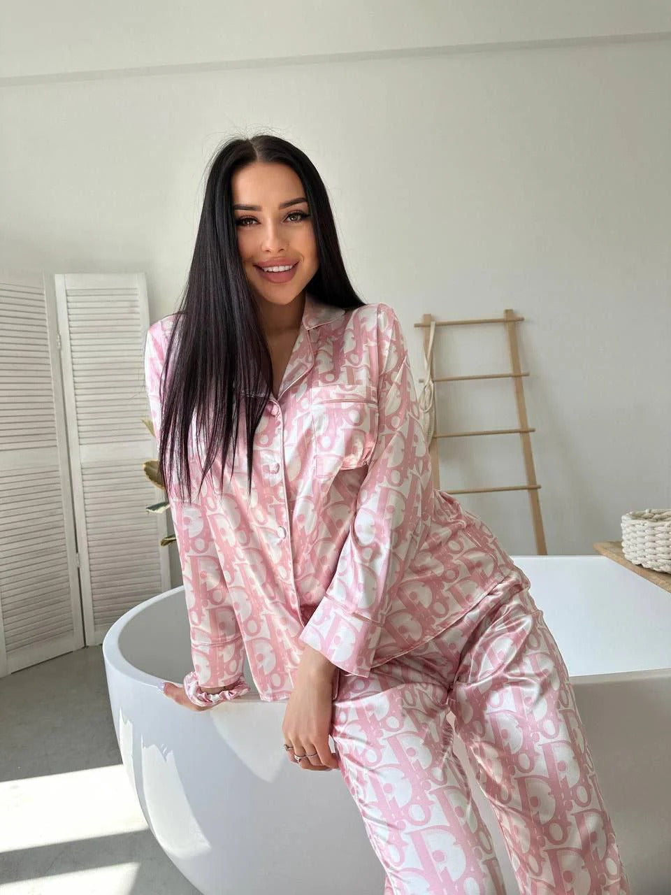 Woman in pink pajamas sitting on a white bathtub in a bright room.