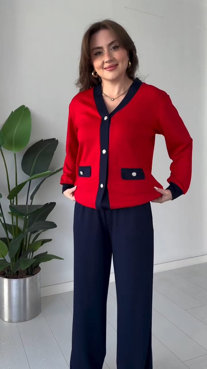 Woman wearing a red cardigan with black accents standing indoors next to a plant.