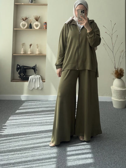 Pakistani women wearing an olive green outfit taking a mirror selfie in a room with a sewing machine and shelves.