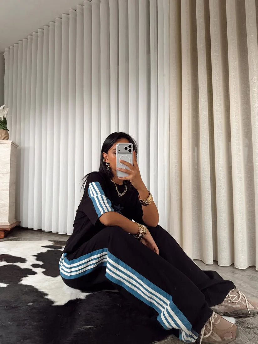 Stylish woman in black oversized co-ord set with striped track pants, sitting indoors and taking a mirror selfie. 01