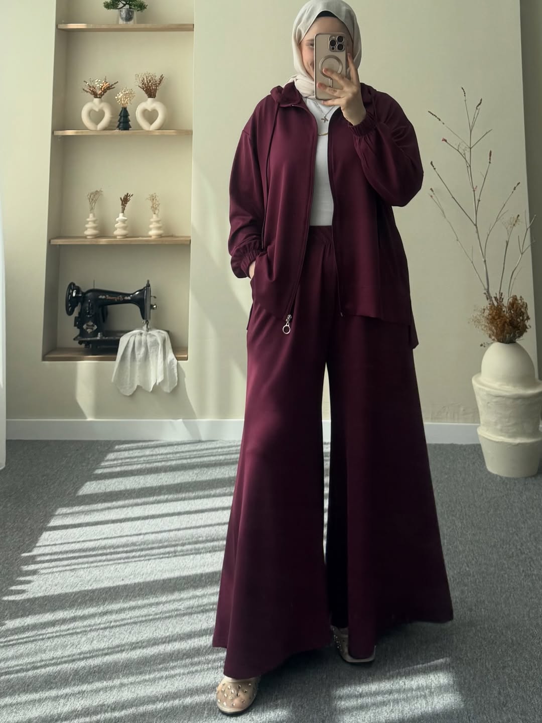 Women wearing a burgundy outfit taking a mirror selfie in a room with a sewing machine and shelves.