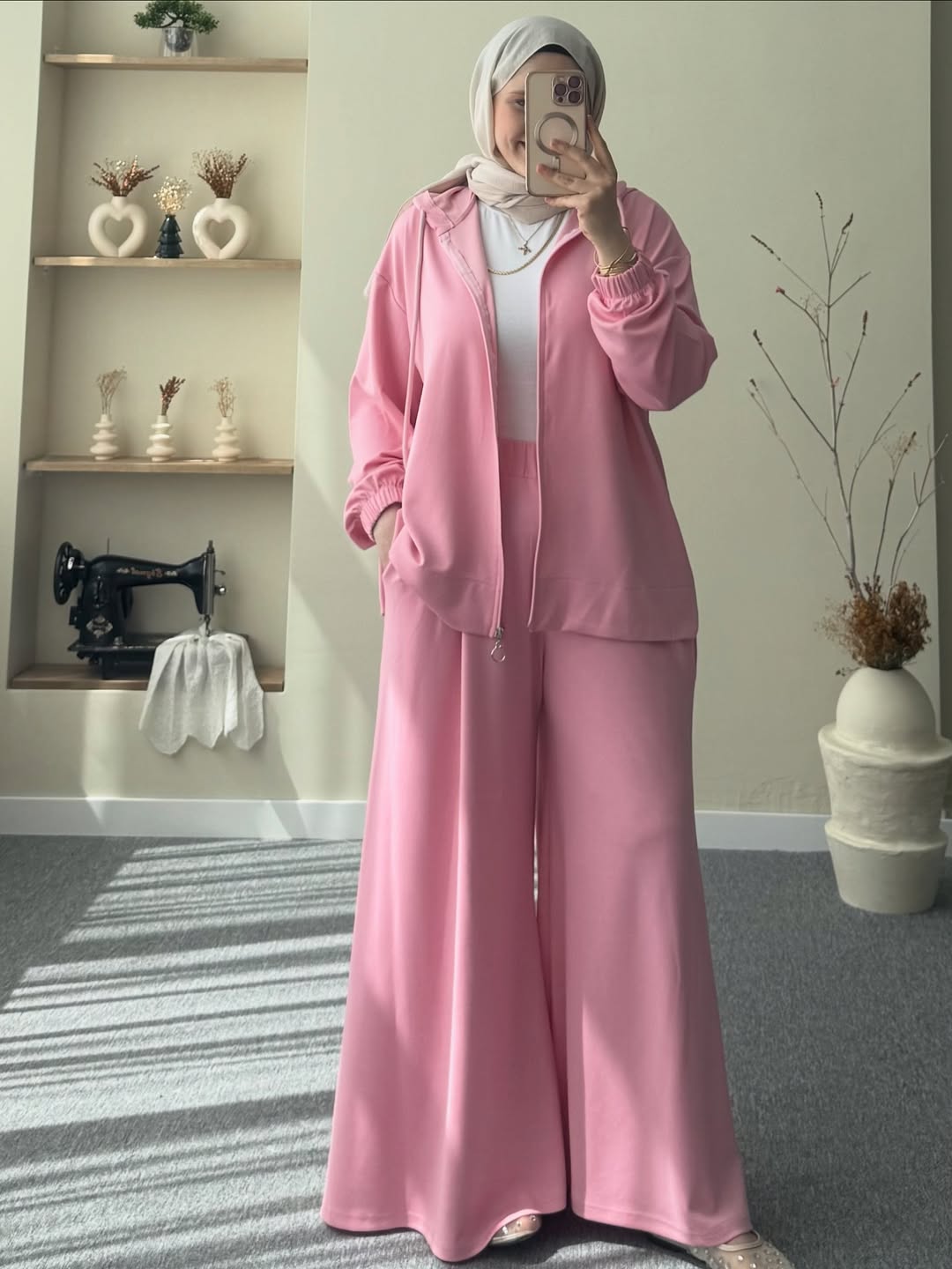 women wearing a pink outfit taking a mirror selfie in a room with a sewing machine and shelves.
